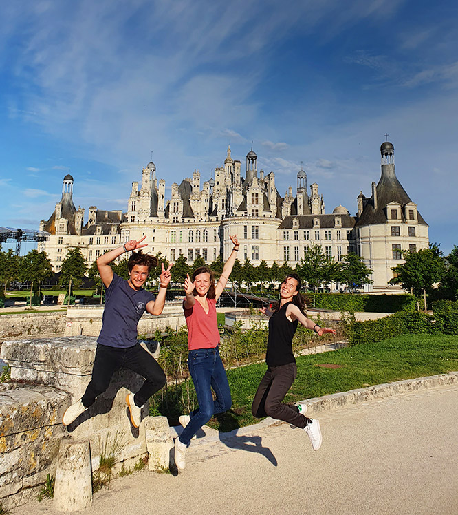 Trois jeunes adultent sautent et sont souriants devant le château de Chambord par temps ensoleillé